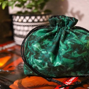 May include: A green fabric dice bag with a black drawstring. The bag has a leafy pattern and is sitting on top of a book.