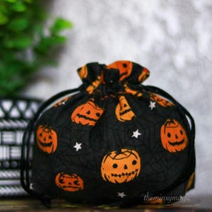 May include: Black drawstring bag with a Halloween theme. The bag is covered in a pattern of orange jack-o'-lantern faces and white stars on a black background.