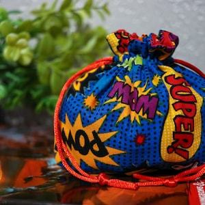 May include: A blue drawstring bag with a red cord. The bag is decorated with a comic book style print featuring yellow, red, and purple speech bubbles with the words "POW", "MOM", and "SUPER!"