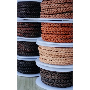 May include: Spools of braided leather cord in brown, tan, and orange colours. The leather cord is arranged in a stack.