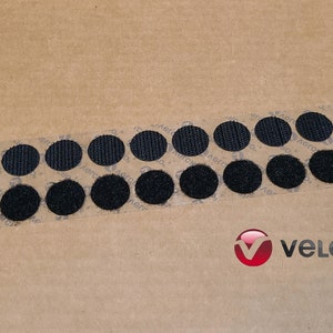 May include: A strip of black hook and loop fasteners, also known as Velcro, on a brown paper background. The strip contains 18 round fasteners.