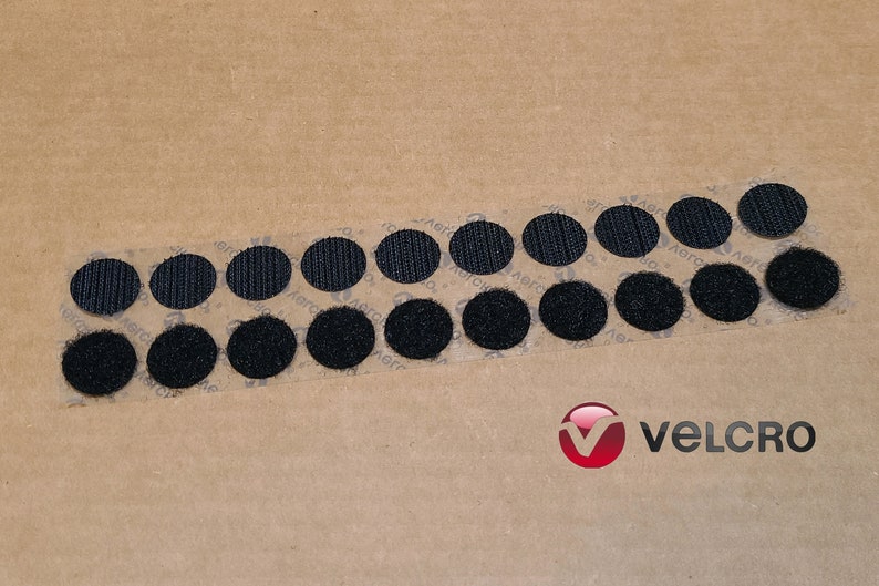 May include: A strip of black hook and loop fasteners, also known as Velcro, on a brown paper background. The strip contains 18 round fasteners.