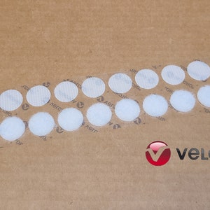 May include: A close-up of a sheet of brown paper with a row of white, round, adhesive Velcro dots. The dots are arranged in a curved line. The Velcro brand logo is visible in the lower right corner of the image.