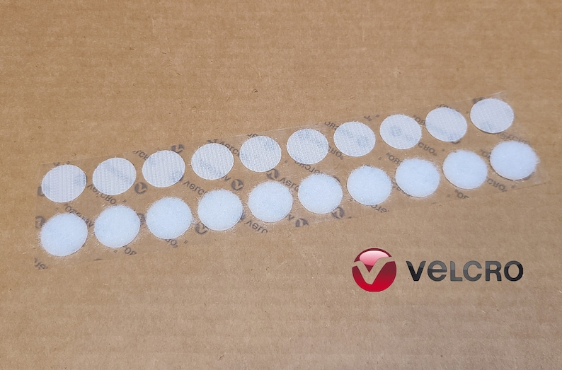 May include: A close-up of a sheet of brown paper with a row of white, round, adhesive Velcro dots. The dots are arranged in a curved line. The Velcro brand logo is visible in the lower right corner of the image.