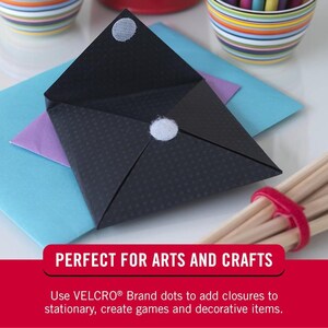 May include: A black envelope with two white adhesive dots on the front. The envelope is folded in half and is sitting on top of a blue, purple, and white envelope. The text "PERFECT FOR ARTS AND CRAFTS" is at the bottom of the image. The text below that reads "Use VELCRO&reg; Brand dots to add closures to stationery, create games and decorative items."