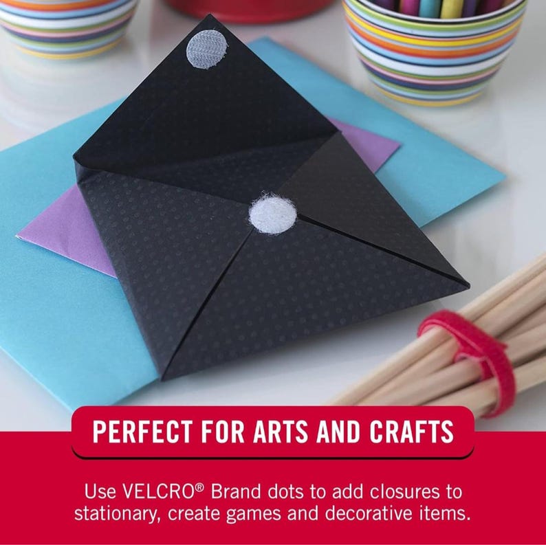 May include: A black envelope with two white adhesive dots on the front. The envelope is folded in half and is sitting on top of a blue, purple, and white envelope. The text "PERFECT FOR ARTS AND CRAFTS" is at the bottom of the image. The text below that reads "Use VELCRO&reg; Brand dots to add closures to stationery, create games and decorative items."
