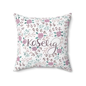 May include: A white cushion with a floral pattern in shades of pink and green. The cushion has the word "koselig" written in brown cursive lettering.