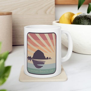 May include: White ceramic mug with a graphic of Haystack Rock at Cannon Beach, Oregon. The graphic features a sunset with orange, pink, and yellow rays of light. The text "Cannon Beach, OR" is printed below the graphic.