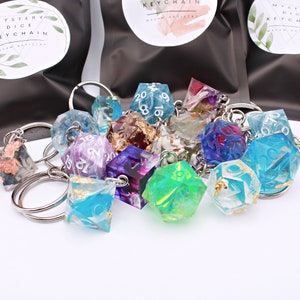 May include: A collection of colorful, resin dice keychains. The dice are various geometric shapes, including d4, d6, d8, d10, d12, and d20. Each die is a different color, with some containing gold leaf or other decorative elements. The keychains are attached to silver-colored metal rings.