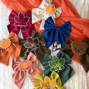 May include: A collection of fabric hair bows in various colours including burgundy, cream, navy blue, orange, brown, green, and mustard yellow. The bows feature embroidered patches of pumpkins, maple leaves, mushrooms, and a teddy bear face.
