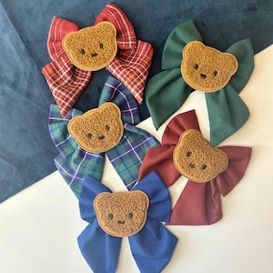 May include: A selection of hair bows in various colours, each adorned with a teddy bear applique. The bows include red plaid, green, blue, and burgundy. The teddy bear appliques are brown with embroidered features.
