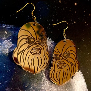 May include: Gold mirrored earrings featuring a detailed outline of Chewbacca's face. The earrings are oval shaped and have a gold hook.
