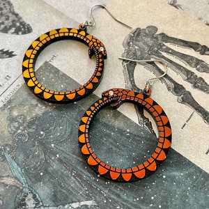 May include: A pair of orange and black snake earrings. The earrings are made of wood and have a snake design. The snake is curled into a circle and has a small head with a tongue sticking out.