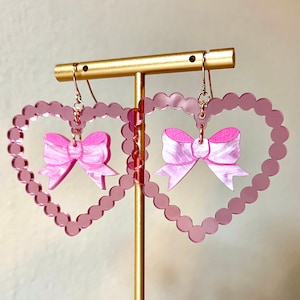 May include: Pink heart-shaped earrings with a pink bow in the center. The earrings are on a gold stand.