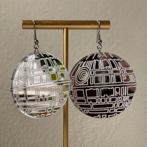 May include: A pair of silver mirrored earrings with a white outline of the Death Star from Star Wars. The earrings are hanging from a gold stand.