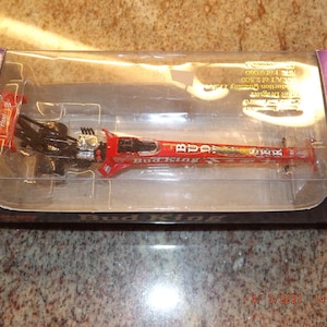 May include: A red, white, and black 1/24 scale diecast model of a Bud King dragster. The model is in a clear plastic display case with a black and purple cardboard box. The box has the words "ACTION" printed on it.