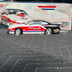 Puede incluir: Modelo a escala de un coche de carreras Dodge, con una combinación de colores blanco, rojo y negro y logotipos de patrocinadores. El coche tiene neumáticos Goodyear y el texto "Johnson Johnson Greenville Oil & Petroleum". La caja muestra el logotipo de Amoco.