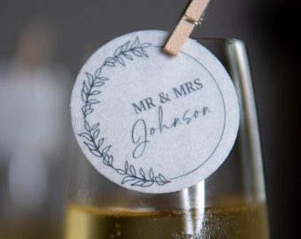 Puede incluir: Un primer plano de una copa de champán con una etiqueta de papel blanco unida al borde. La etiqueta tiene un diseño de corona negra y el texto "MR & MRS Johnson".