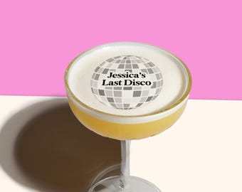 May include: A cocktail in a stemmed glass with a yellow liquid and white foam. The foam has a gray and white disco ball graphic with the text "Jessica's Last Disco". The background is pink and white.
