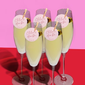 Puede incluir: Cinco copas de champ&aacute;n con etiquetas circulares rosas y blancas que dicen "Chloe's Hen Do 24.05.23" sujetas a los tallos con pinzas de madera.