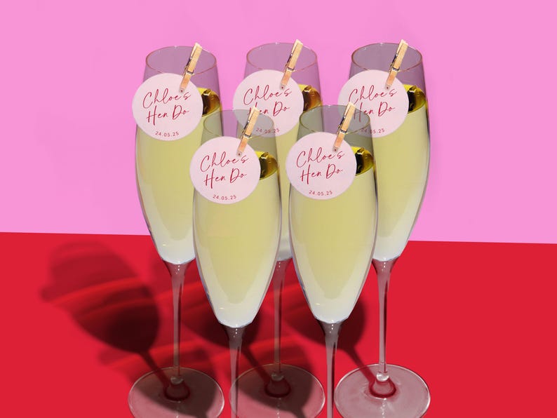 Puede incluir: Cinco copas de champ&aacute;n con etiquetas circulares rosas y blancas que dicen "Chloe's Hen Do 24.05.23" sujetas a los tallos con pinzas de madera.