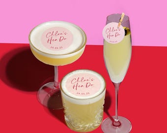 May include: Two cocktails with pink and white round stickers that say "Chloe's Hen Do 24.05.25" on them. The cocktails are in clear glasses and are on a red surface.