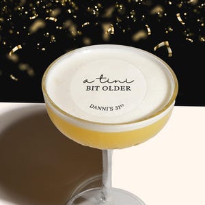 May include: A cocktail with a white foam top and a round sticker that says "a  tini bit older" and "Danni's 31st" in black text.