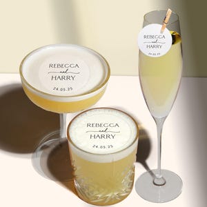 May include: Two clear glass cocktail glasses with a white frosted rim. The glasses are filled with a yellow liquid and have a white sticker on the rim that says "Rebecca and Harry 24.05.25".