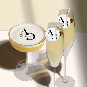 May include: Three clear glass cocktail glasses with a white circle sticker on each. The stickers have the letters "A" and "D" in black with the text "The Morris" in black cursive underneath.