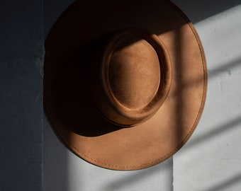 May include: A brown suede fedora hat with a wide brim and a stitched edge.