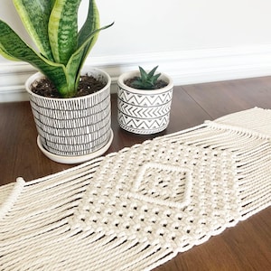 May include: A white macrame table runner with a diamond pattern. The runner is on a wooden table with two potted plants, one with green leaves and the other with a succulent.