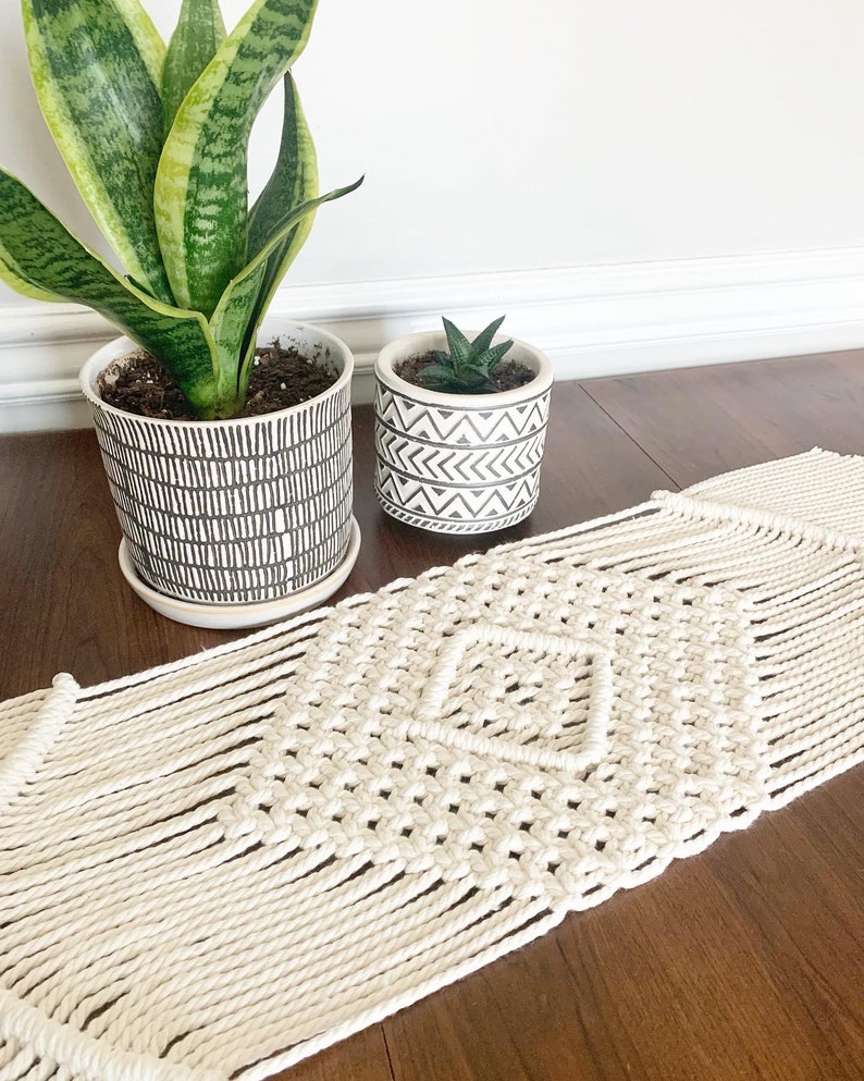 以下が含まれることがあります： ダイヤモンド模様の白いマクラメテーブルランナー。ランナーは木製のテーブルの上にあり、緑の葉の植物と多肉植物の2つの鉢植えがあります。