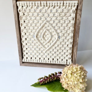 May include: A white macrame wall hanging with a diamond pattern in a wooden frame. The macrame is made with thick cotton cord and has a natural, bohemian style.