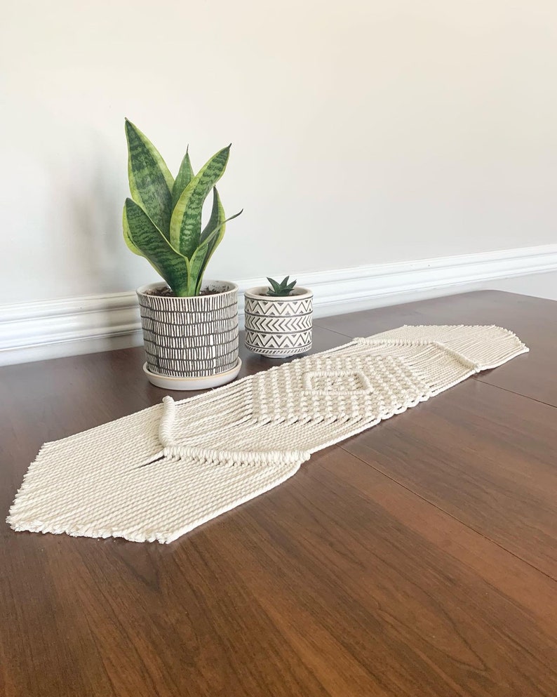 以下が含まれることがあります： ダイヤモンド模様の白いマクラメテーブルランナー。ランナーは木製のテーブルの上にあり、蛇の植物と多肉植物の2つの鉢植えがあります。
