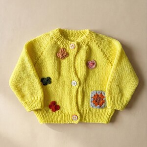 Puede incluir: Un cárdigan de punto amarillo brillante con mangas largas, cuello redondo y tres botones con diseños de caras sonrientes. La prenda está decorada con pequeñas aplicaciones de ganchillo de colores, incluyendo flores, mariposas y un parche cuadrado.