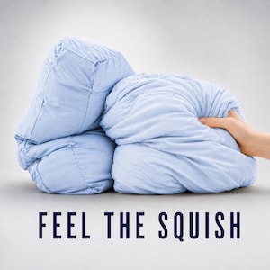 May include: A light blue quilted blanket with a soft, folded texture. The fabric appears pliable and plush, with visible stitching lines creating a grid pattern on the left side. The text "FEEL THE SQUISH" is printed in dark blue capital letters below.