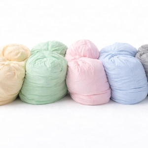 May include: A collection of seven beanbag chairs in various pastel colors, including yellow, green, pink, blue, and gray. The beanbags are arranged in a row against a white background, showcasing their soft, rounded shapes.