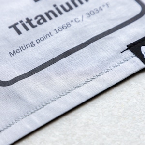 Puede incluir: Primer plano de una bolsa de tela gris con costuras negras. La bolsa tiene una etiqueta que dice "Titanio Punto de fusión 1668°C / 3034°F". Una etiqueta negra con el texto "elefanti  STUFF" está unida a la bolsa.