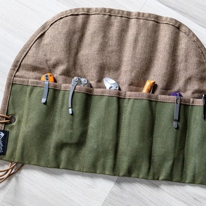 Puede incluir: Un rollo de lona verde con seis bolsillos para guardar herramientas. El rollo tiene una solapa marrón y una correa de cuero con la palabra "Griffey" impresa.