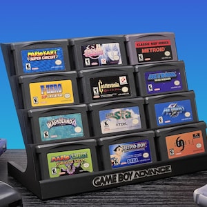 May include: A black Game Boy Advance cartridge display stand holding various game cartridges. The cartridges feature titles like "Mario Kart: Super Circuit", "Metroid", and "Astro Boy". The stand is labeled "Game Boy Advance".
