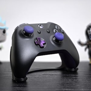 May include: Black Xbox controller with purple accents, including thumbsticks and a directional pad. Two figurines are in the background, one of Rick from Rick and Morty and another of a character in a hooded cloak.
