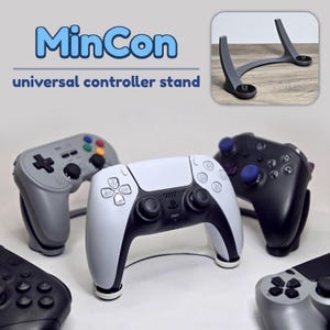 Könnte beinhalten: Ein MinCon Universal-Controller-Ständer in einem oberen Einschubbild, auf dem ein weiß-schwarzer Game-Controller ruht. Weitere graue und schwarze Game-Controller sind um den Ständer auf einer weißen Oberfläche angeordnet und zeigen verschiedene Layouts.