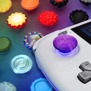 Puede incluir: Una consola de juegos portátil blanca con una tapa de joystick morada texturizada y un pad direccional gris. Alrededor de la consola hay varias cubiertas de joystick circulares y coloridas en tonos amarillo, naranja, rojo, negro, verde y azul.