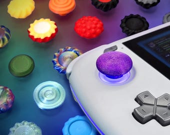 Può includere: Una console di gioco portatile bianca con un cappuccio per joystick viola testurizzato e un pad direzionale grigio. Attorno alla console si trovano vari copri-joystick circolari e colorati in tonalità giallo, arancione, rosso, nero, verde e blu.