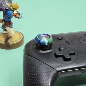 May include: A black video game controller featuring a circular, iridescent thumbstick cap with a metallic, spiral-grooved texture. The controller has a textured grip and a directional pad. A small figurine of a character in a blue tunic stands nearby.