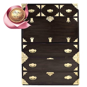 May include: A dark wood chest of drawers with brass hardware and decorative accents. The chest has six drawers, including a small top drawer with a drop-down front.