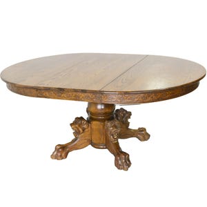 May include: A large, oval, wooden dining table with a dark brown finish. The table has a pedestal base with three lion's paw feet. The table is made of solid wood and has a beautiful grain pattern.