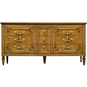 May include: A wooden dresser featuring a rectangular frame with a light brown finish. It includes six wide drawers on the sides and three narrow central drawers, all adorned with gold-toned metal hardware, including curved handles and small knobs.