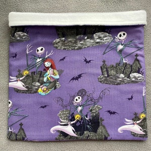 May include: Purple fabric pouch with a white fleece lining. The pouch features a repeating pattern of Jack Skellington and Sally from the Nightmare Before Christmas movie. The characters are depicted in various scenes from the movie, including Jack Skellington standing on a hilltop and Sally sitting on a cloud.
