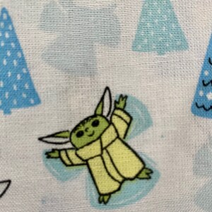 May include: A green and yellow cartoon character with large ears and a robe, making a snow angel on a white background with blue and white Christmas trees.